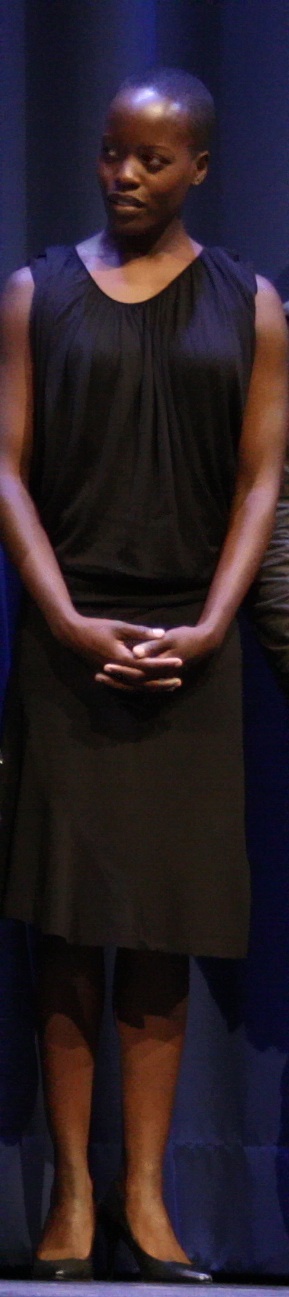 Florence Kasumba in einem langen schwarzen Trägerkleid. Sie hat die Hände vor dem Körper ineinander verschränkt.