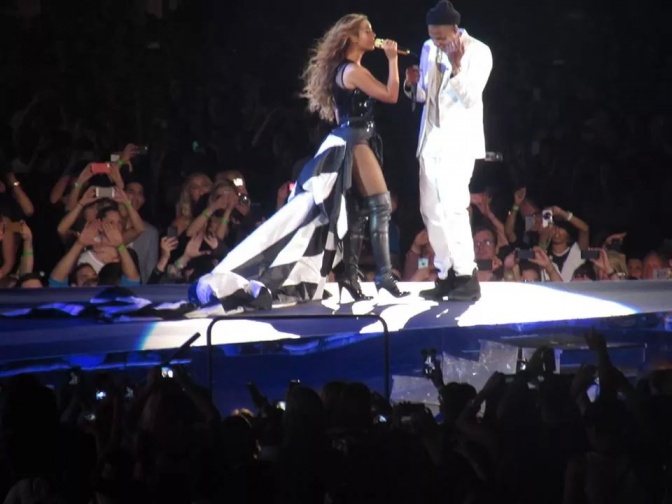 Jay-Z und Beyoncé zusammen auf der Bühne. Sie trägt ein schwarzes, er ein weißes Outfit. Beide singen in ein Mikrophon. ImHintergrund sieht man die Zuhörerinnen und Zuhörer.