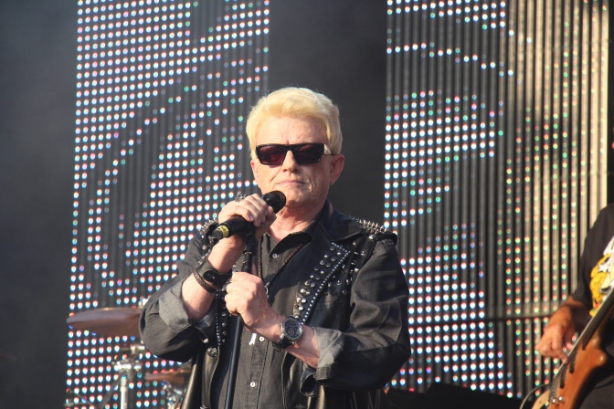 Heino steht auf der Bühne und singt in ein Mikrophon. Er trägt eine Lederjacke und eine große, schwarze Sonnenbrille.