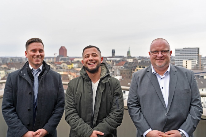 3 Männer stehen lächelnd nebeneinander. Im Hintergrund sieht man die Skyline der Stadt Duisburg.