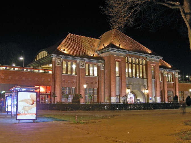 Ein großes Bahnhofsgebäude mit dem Schriftzug Mundsburg über dem Eingangsportal. Links vorne im Bild sind 2 Bushaltestellen mit Leuchtreklame zu sehen.
