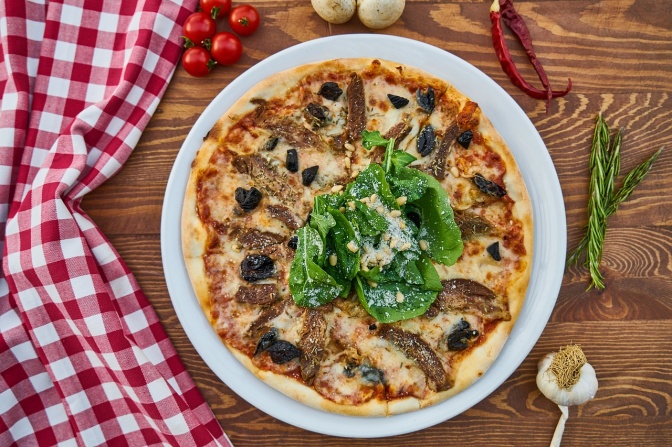 Eine Pizza, belegt mit Käse, Sardellen, Oliven und frischem Basilikum. Der Pizzateller steht auf einem Holztisch, der zur Hälfte von einer karierten Tischdecke bedeckt ist.