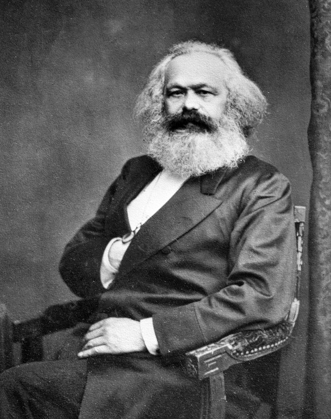 Ein altes schwarz-weiß-Foto von Karl Marx in sitzender Position. Er hat lange, weiße Haare und einen langen, weißen Bart.