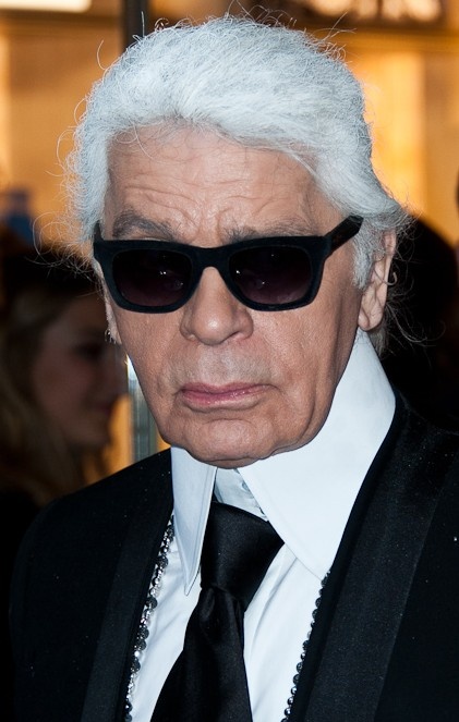 Karl Lagerfeld mit weißen, zum Zopf gebundenen Haaren und schwarzer Sonnenbrille. Er trägt Anzug und Krawatte.