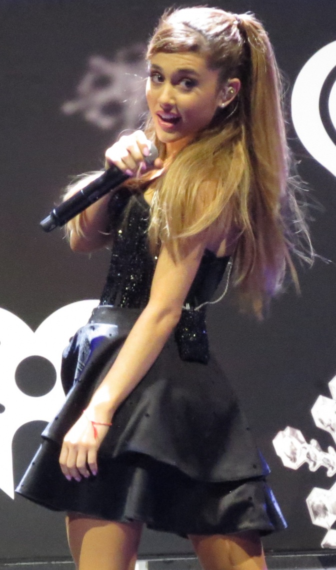 Ariana Grande in einem kurzen schwarzen Kleid. Sie hat einen Pferdeschwanz und singt in ein Mikrophon.