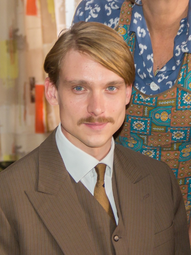 Daniel Sträßer in einem braunen Anzug mit passender Krawatte. Er trägt einen Seitenscheitel und einen blonden Schnurrbart.