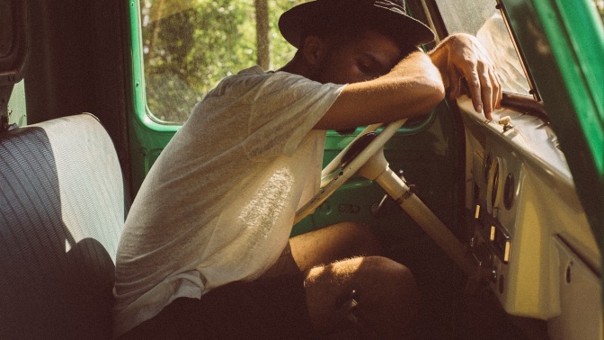 Ein Mann mit einem Strohhut auf dem Kopf schläft, über das Lenkrad eines Busses gebeugt.
