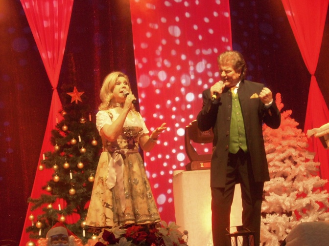 Marianne und Michael auf einer Bühne mit viel Dekoration und künstlichen Bäumen. Die beiden singen in zwei Mikrophone. Sie tragen Tracht.