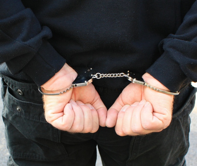 Eine Person in einer dunklen Jacke mit auf den Rücken gefesselten Händen in Handschellen.