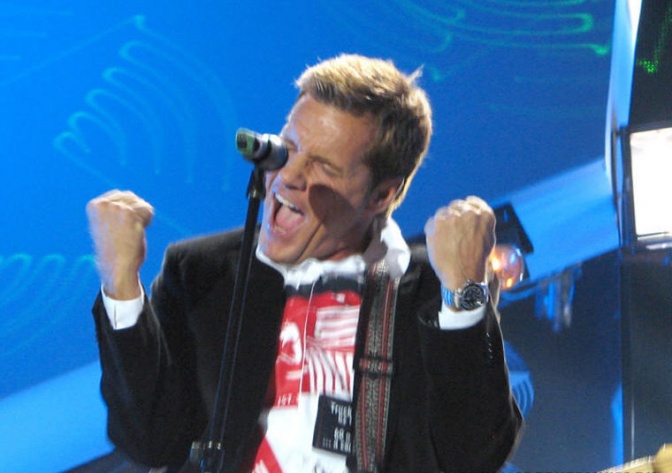 Dieter Bohlen steht auf der Bühne vor einem Mikrophon. Er streckt beide Fäuste in Siegerpose in die Höhe. Er trägt ein gemustertes Shirt mit schwarzem Sakko.