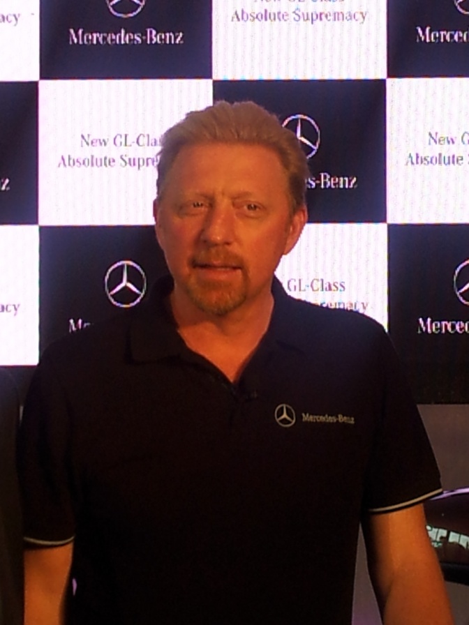 Boris Becker steht in einem schwarzen T-Shirt vor einer Logowand mit verschiedenen Markenschriftzügen. Unter anderem dem Logo von Mercedes Benz.