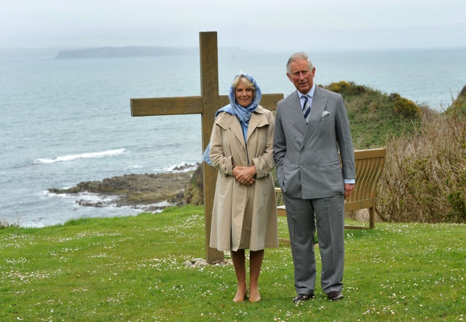 Prinz Charles und seine Frau Camilla stehen auf einer Wiese. Hinter ihnen sieht man ein großes Holzkreuz und das Meer. Prinz Charles trägt einen Anzug, Camilla einen Mantel und Kopftuch.