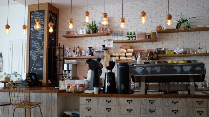 Der Innenraum eines Cafés mit einer Profi-Kaffeemaschine.  Die Getränkeauswahl ist an eine Tafel geschrieben. Ein Mann steht hinter dem Tresen.