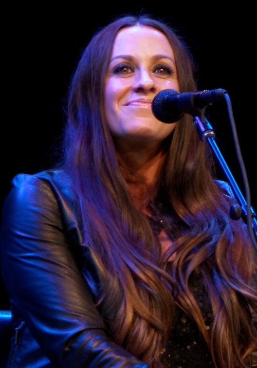 Alanis Morissette mit langen, braunen gewellten Haaren. Sie trägt eine Lederjacke und sitzt lächelnd vor einem Mikrophon.