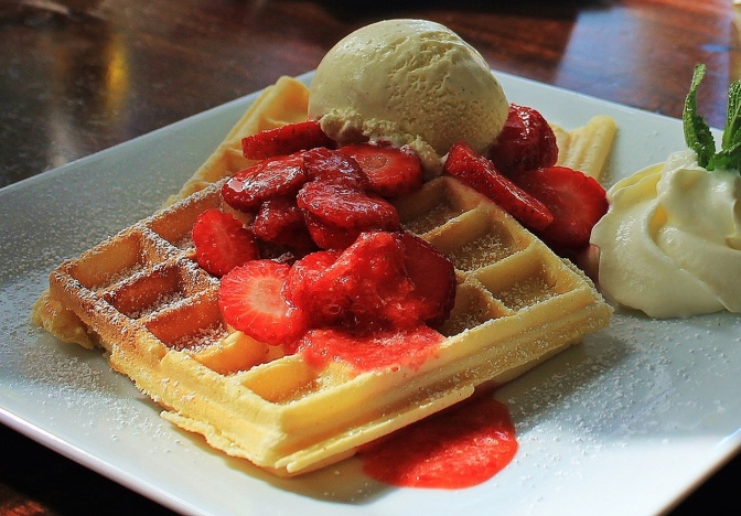 Eine quadratische Waffel mit Erdbeeren und Vanilleeis auf einem weißen, eckigen Teller.