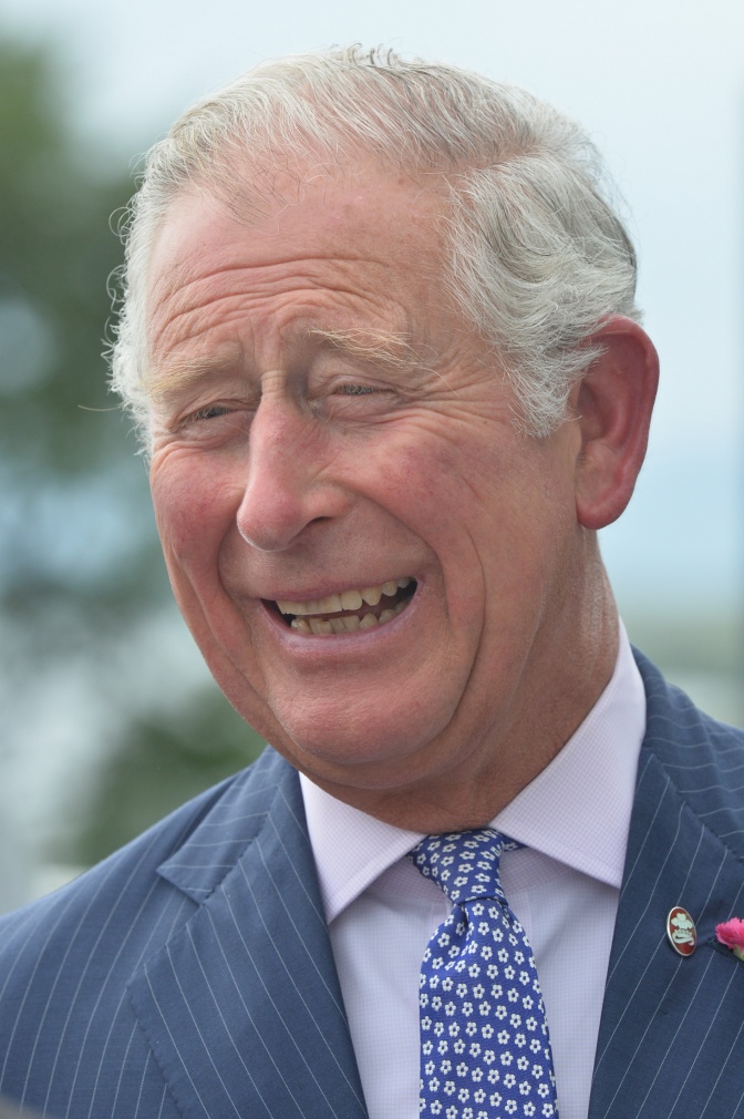 Prinz Charles in Anzug und Krawatte. Er lacht. Er hat graue Haare und hat einige lange Strähnen über die Stirnglatze frisiert.
