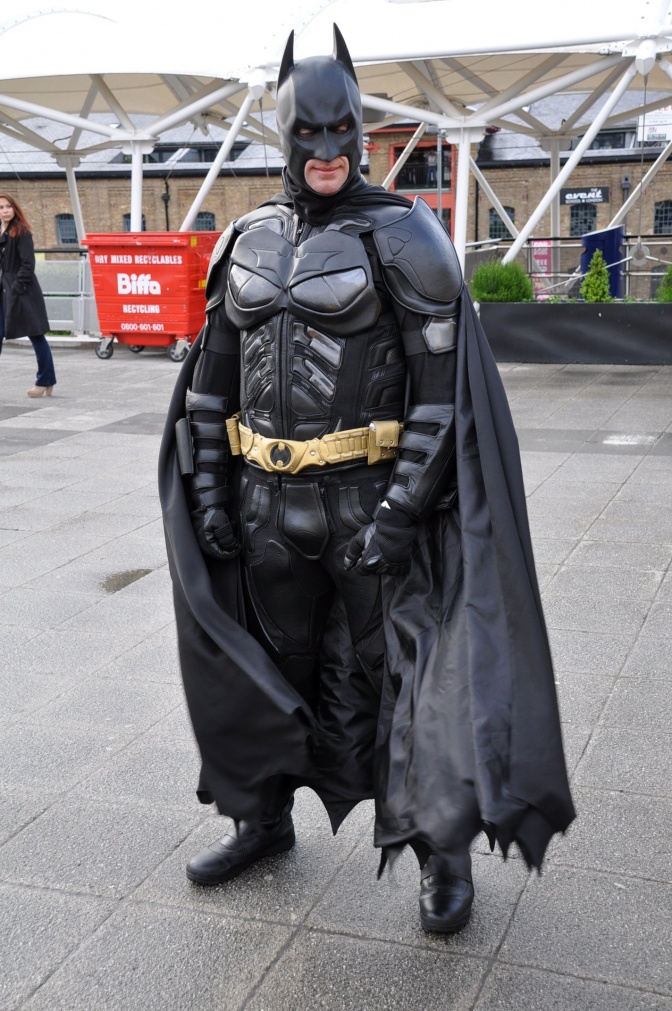 Ein Mann in einem Batman-Kostüm steht vor einem Pavillon und einem Müllcontainer. Das Kostüm ist schwarz und erinnert an eine Fledermaus.