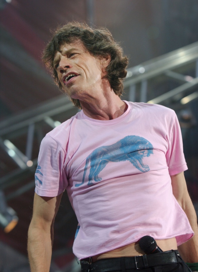 Mick Jagger auf der Bühne. Er trägt ein enges T-Shirt mit einem Löwen auf der Brust und hat sich das Mikrophon in den Hosenbund gesteckt.