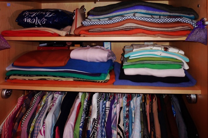 Das Innere eines Kleiderschranks mit gefalteten und gestapelten Kleidungsstücken auf einem Bord und Kleidungsstücken, die an Kleiderbügeln hängen.