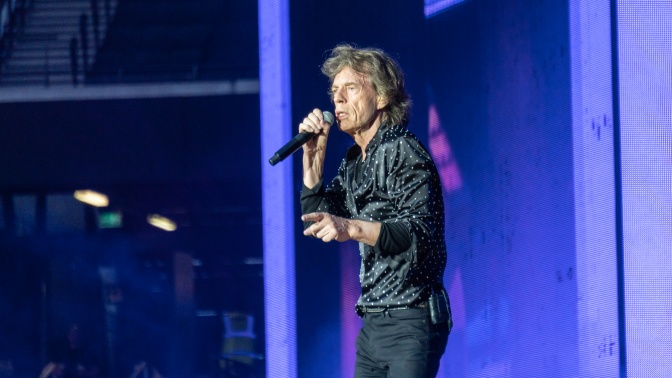 Mick Jagger auf der Bühne. Er trägt ein schwarzes Outfit und singt in ein Mikrophon.