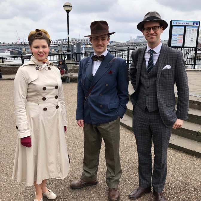 2 Männer und eine Frau in Vintage-Kleidung und mit Hüten. Im Hintergrund sieht man die Themse und Brücken.