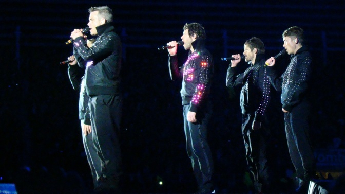 Take That stehen in Formation auf der Bühne und singen in Handmikrophone. Sie tragen schwarze Jacken mit leuchtenden LEDs.