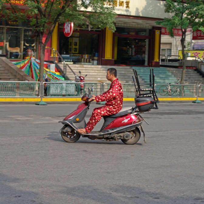Ein asiatischer Mann im Schlafanzug auf einem Motorroller. Er transportiert einen Stuhl auf dem Roller.
