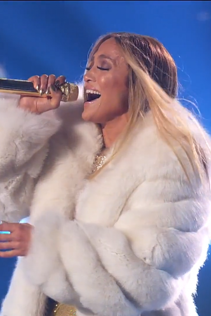 Jennifer Lopez mit langen, blonden offenen Haaren. Sie trägt eine weiße Pelzjacke und singt in ein Handmikrophon.