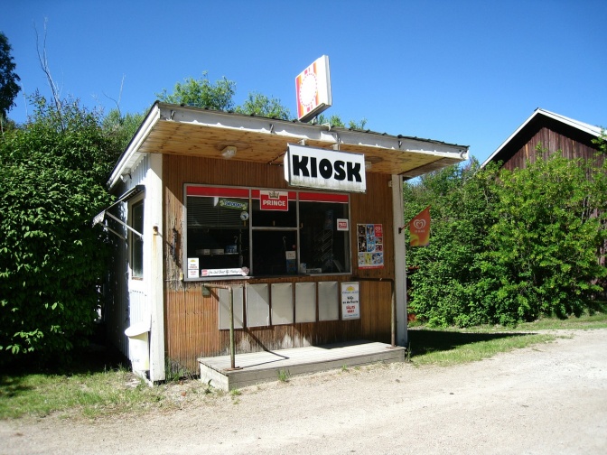 Ein Kiosk in einer kleinen Bretterbude. Die Fassade ist mit Werbung beklebt für Eis, Schokoriegel und Zigaretten.