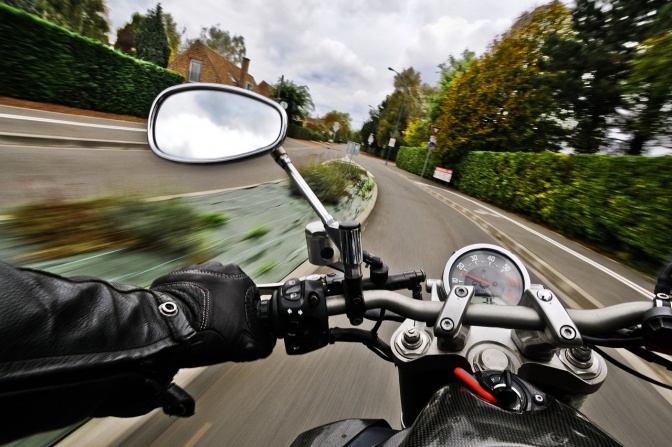 Ein Motorrad fährt über eine von Bäumen umstandene Straße. Man sieht nur Lederhandschuhe, die den Lenker halten.