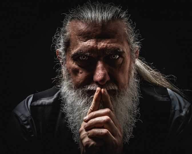 Das Porträt eines älteren Mannes mit langen grauen Haaren. Er hält beide Finger an die Nase.