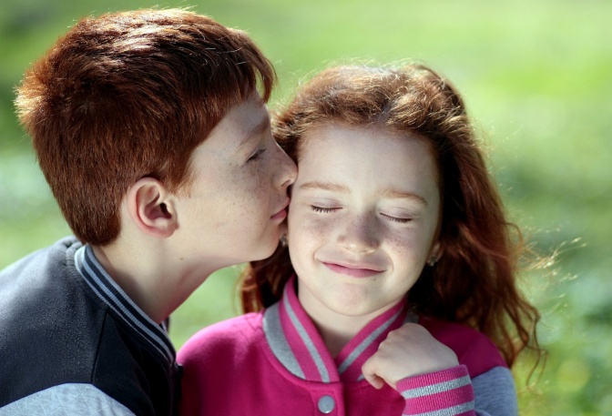 Ein Junge küsste ein Mädchen auf die Schläfe. Sie schließt dabei die Augen. Beide haben rote Haare. Im Hintergrund sieht man eine Wiese.