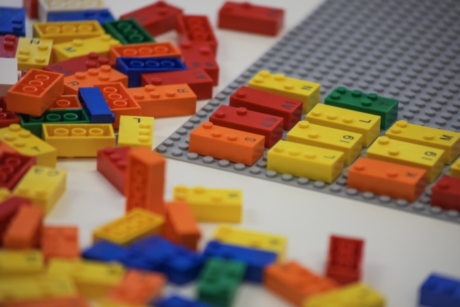 Legosteine in verschiedenen Farben auf einer Lego-Platte. Sie sind in Brailleschrift beschriftet, auf jedem Stein sieht man auch den gedruckten Buchstaben in Schwarzschrift dazu.