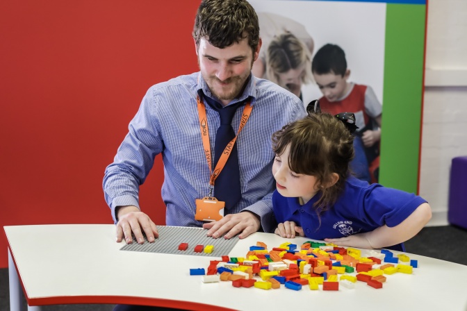 Ein Mann mit Krawatte und ein Mädchen probieren die Legosteine mit Brailleschrift zusammen aus. Sie befühlen sie mit den Händen. Dabei stehen sie nebeneinander an einem Stehtisch. Im Hintergrund sind weitere Personen zu sehen.