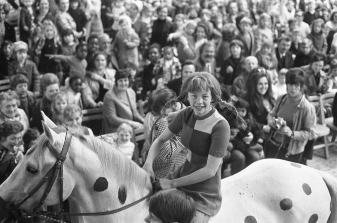 Ein schwarz-weiß-Foto von Inger Nilsson mit abstehenden geflochtenen Zöpfen im Kostüm als Pippi Langstrumpf. Sie reitet auf einem Pferd.