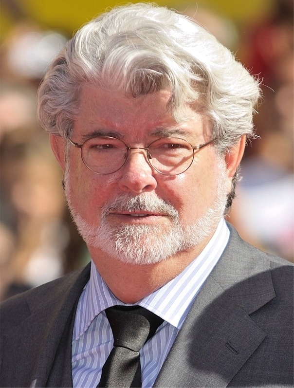 George Lucas mit gewellten Haaren und in Anzug und Krawatte. Er trägt eine schmale Brille.