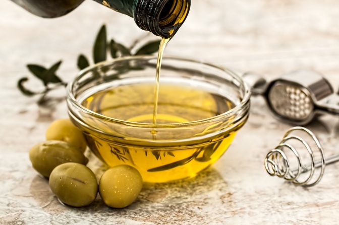 Eine kleines Glasschale, in die Olivenöl gegossen wird. Daneben liegen grüne Oliven.
