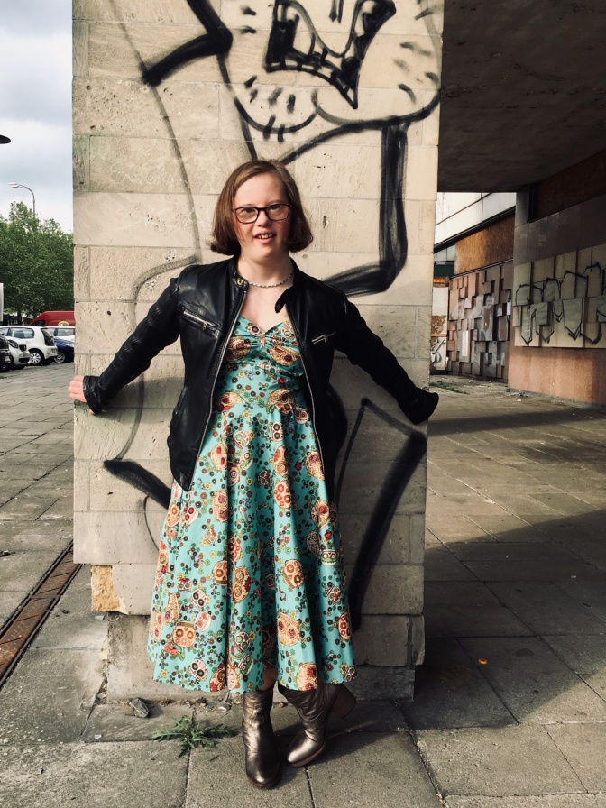 Natalie Dedreux steht in einem gemusterten Kleid mit Lederjacke vor einer Säule mit Graffitis.