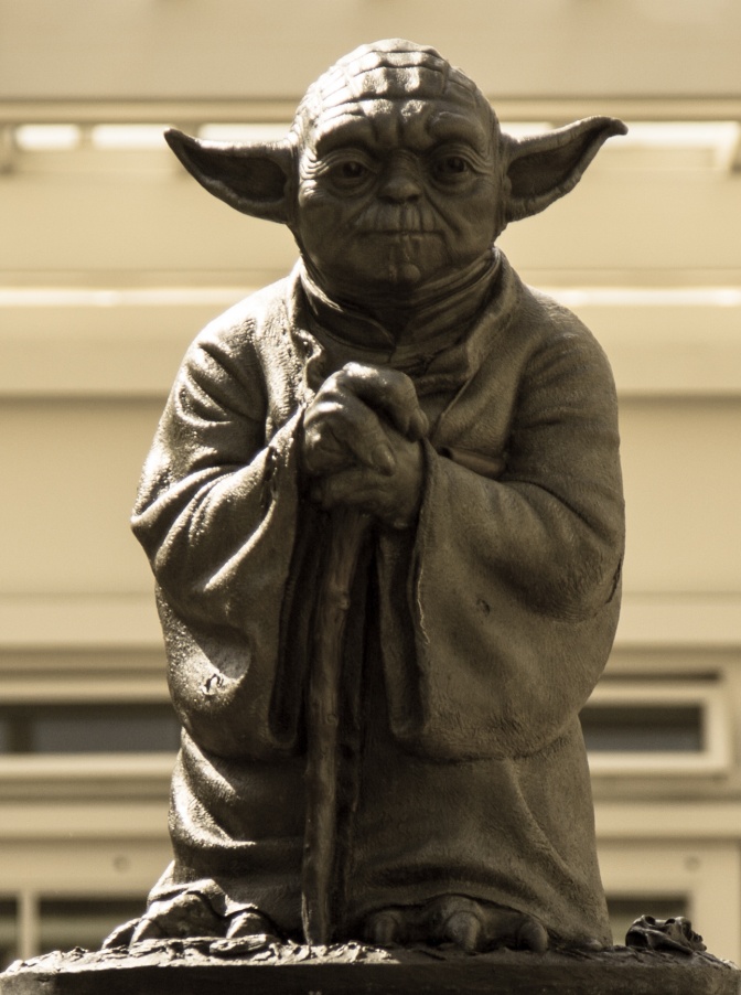 Eine kleine Statue von Yoda mit seitlich abstehenden Ohren. Im Hintergrund sieht man eine Treppe.