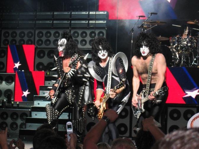 Die Band KISS auf der Bühne. Sie tragen maskenhaftes Make-up. Einer der Musiker streckt die Zunge raus.