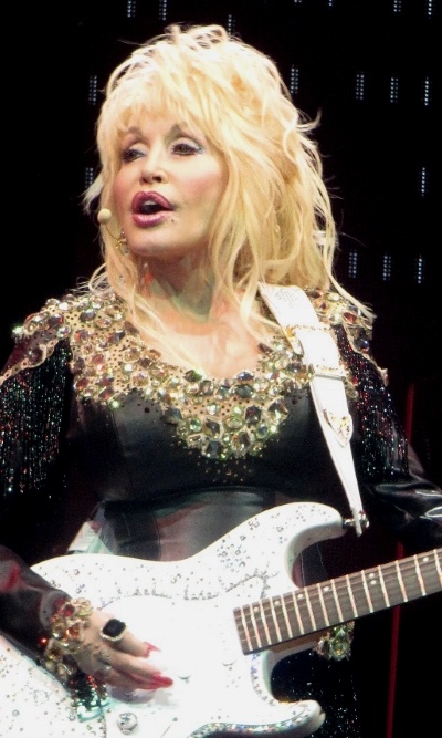 Dolly Parton mit blonden langen Haaren und einer weißen E-Gitarre