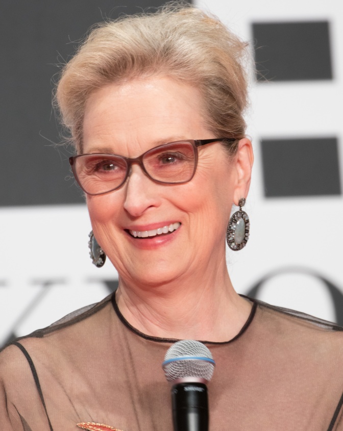 Meryl Streep in einem transparenten Kleid, mit hochgesteckten Haaren und mit hängenden Ohrringen. Sie lächelt und steht vor einem Mikrophon.