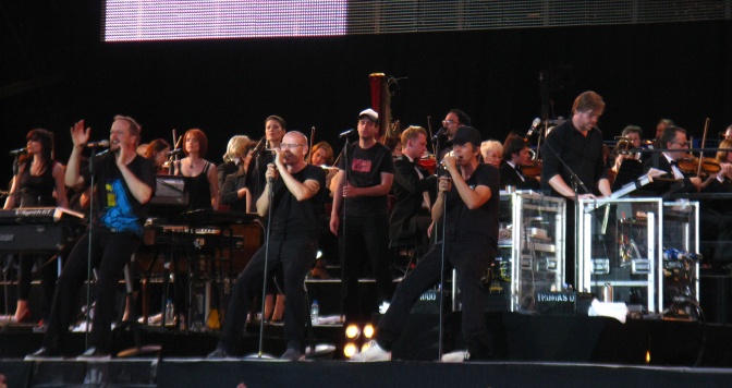 Die Fantastischen Vier in schwarzer Kleidung auf der Bühne. Sie stehen vor Standmikrophonen. Hinter ihnen spielt ein Orchester.