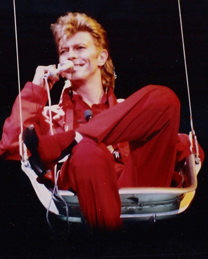 David Bowie mit Vokuhila-Haarschnitt. Er sitzt in einer Schaukel und singt in ein Mikrophon.