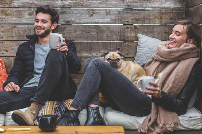 Ein Mann und eine Frau sitzen, zusammen mit einem Mops, auf Kissen. Sie trinken Kaffee.