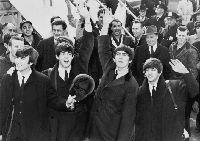 Ein schwarz-weiß-Foto der Band The Beatles. Sie stehen nebeneinander, tragen Anzug, Krawatte und einen schwarzen Mantel. Im Hintergrund sieht man Fans.