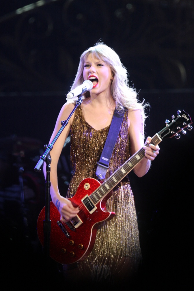 Taylor Swift singt in ein Stand-Mikro und spielt E-Gitarre. Sie hat lange, gewellte blonde Haare.