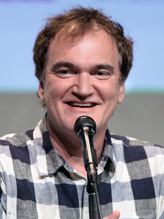 Quentin Tarantino in einem karierten Hemd und mit Geheimratsecken. Er lächelt und spricht in ein Mikrophon.