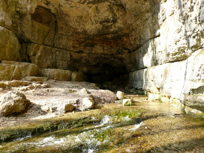 Ein Höhleneingang aus grauem Gestein. Am Boden fließt niedriges Wasser.