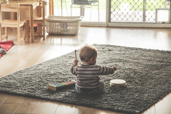 Ein Baby sitzt min dem Rücken zur Kamera auf einem Teppich und spielt ein Xylophon und eine kleine Trommel.
